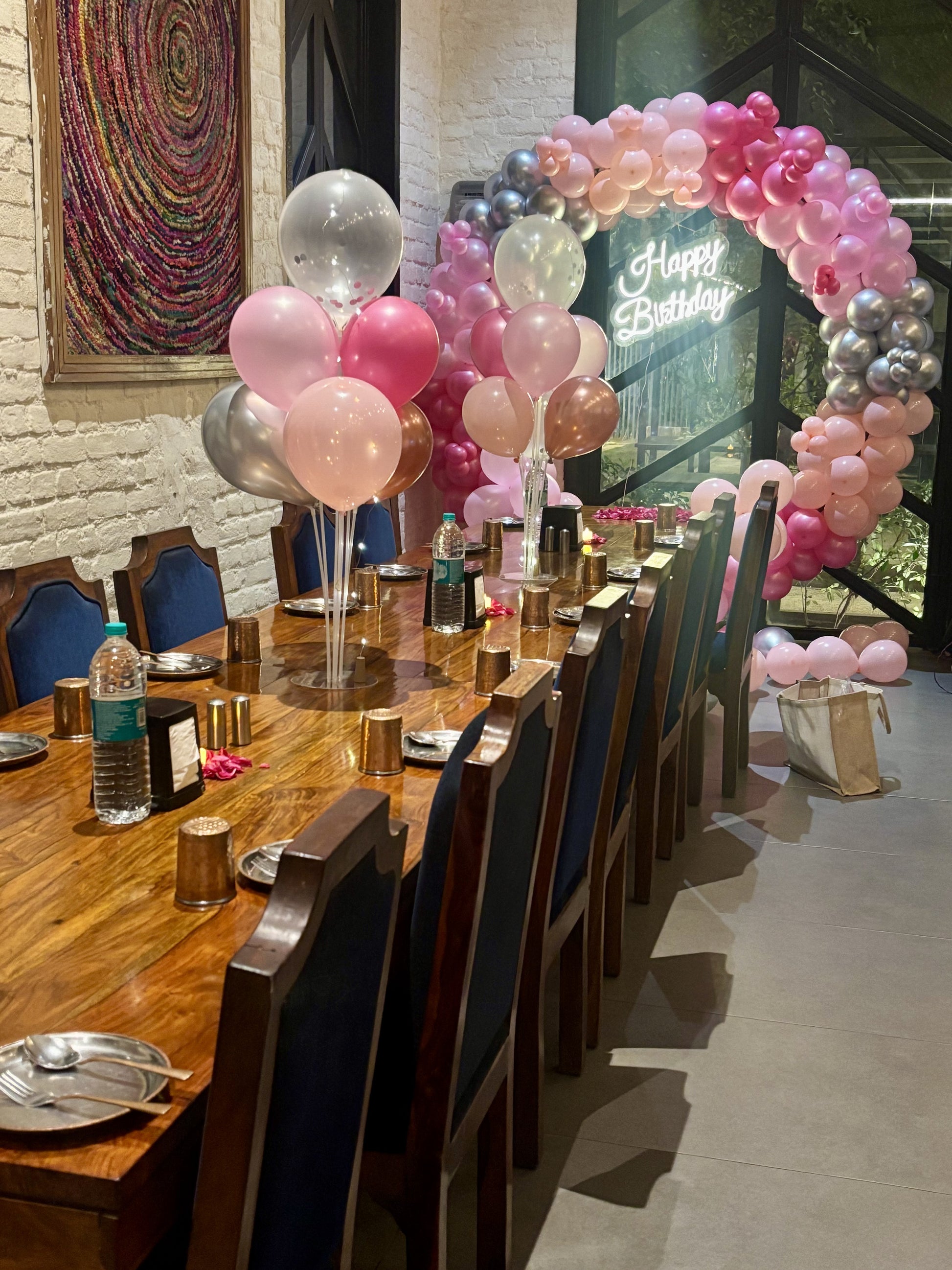 Intimate birthday dinner setup in Jaipur by Shava Moments, featuring a pink and silver balloon arch, neon Happy Birthday sign, and elegant table decorations with rose petals