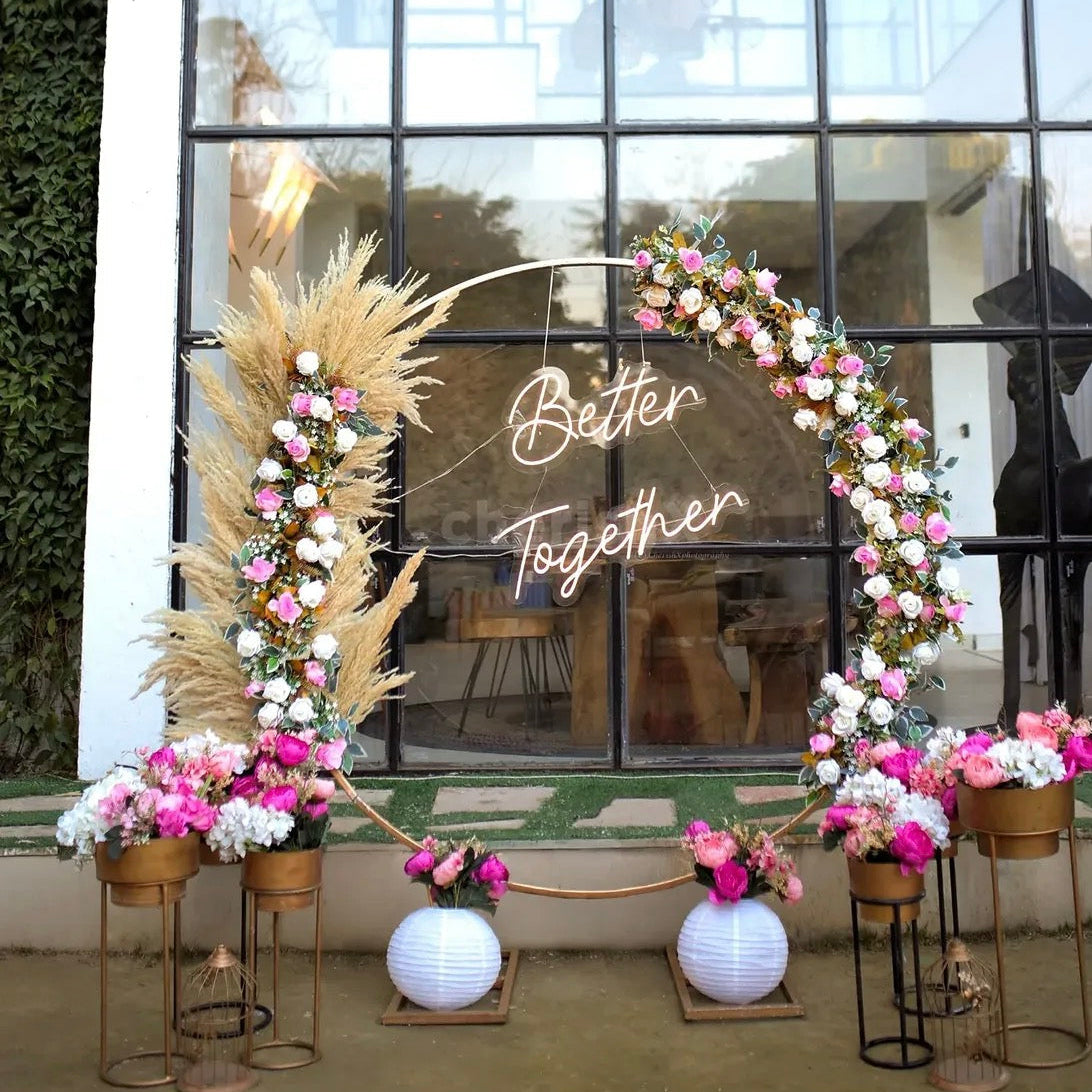 Minimalist circular gold ring backdrop in Jaipur with pampas grass, pink roses, and a Better Together neon sign by Shava Moments.