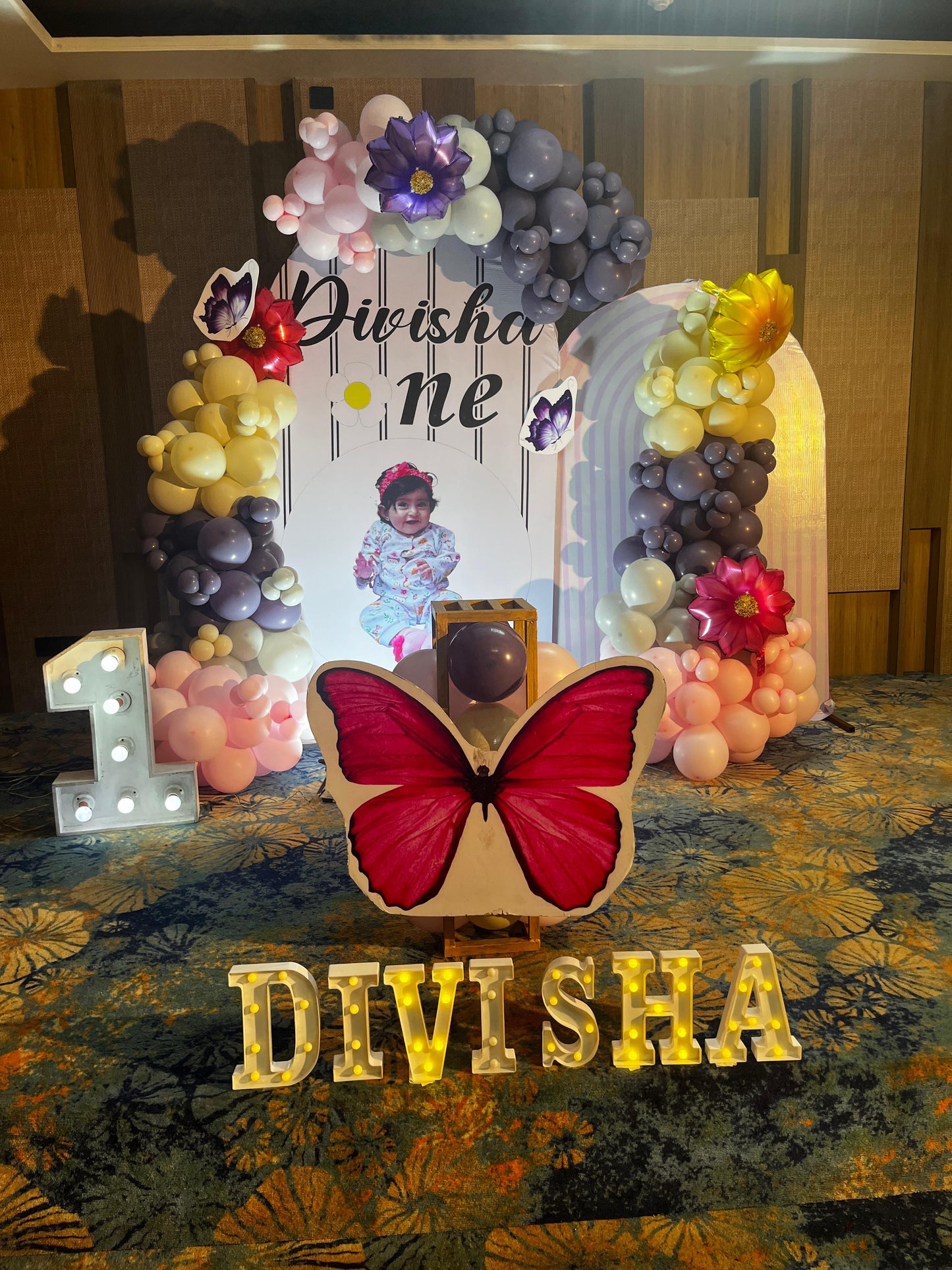 Personalized butterfly and floral 1st birthday backdrop in Jaipur with purple and pink balloons and lighted name sign by Shava Moments.