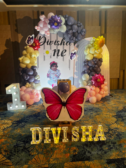 Personalized butterfly and floral 1st birthday backdrop in Jaipur with purple and pink balloons and lighted name sign by Shava Moments.