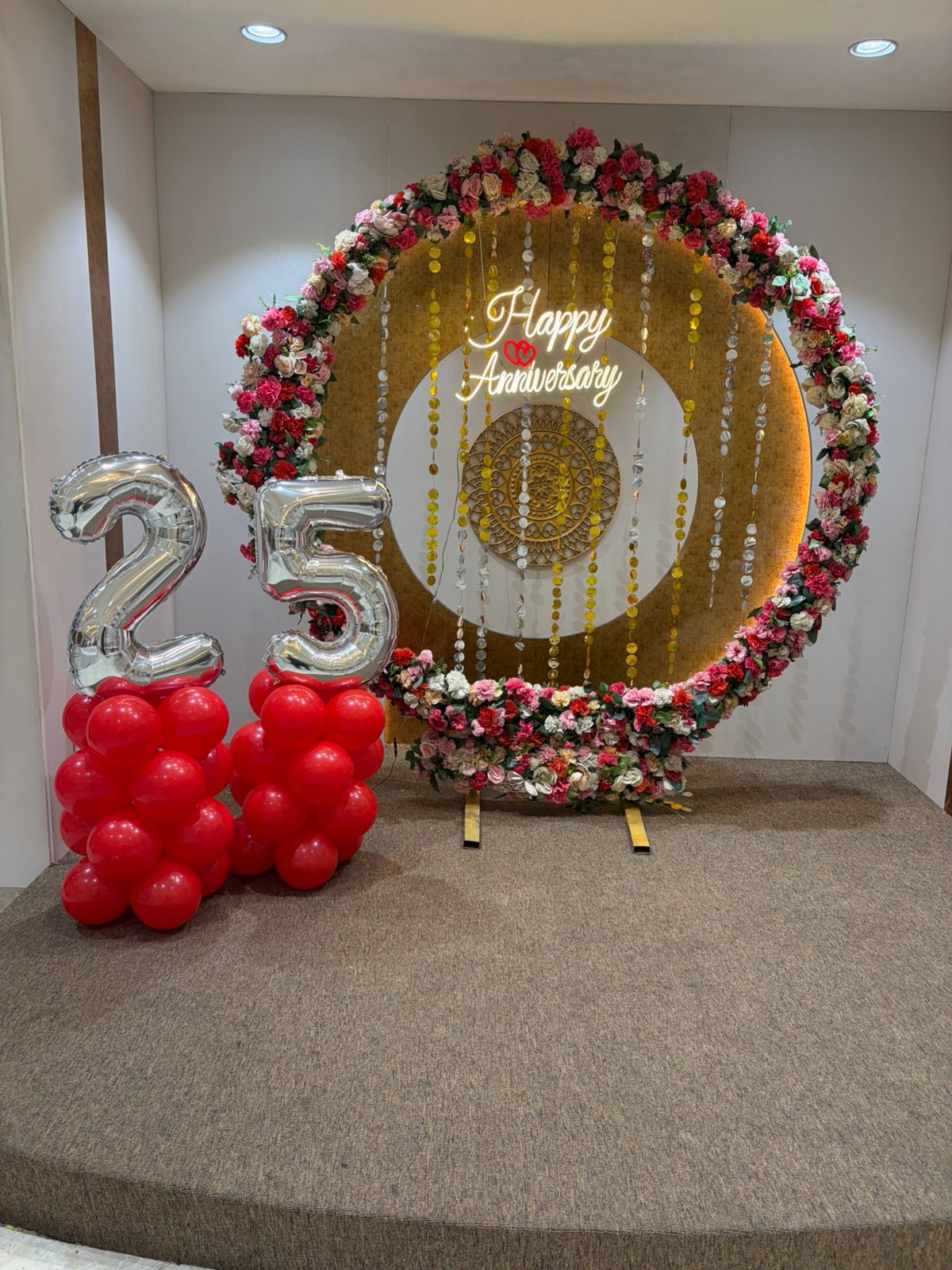 Circular floral ring 25th anniversary backdrop in Jaipur with a neon sign and red balloon pillars by Shava Moments.