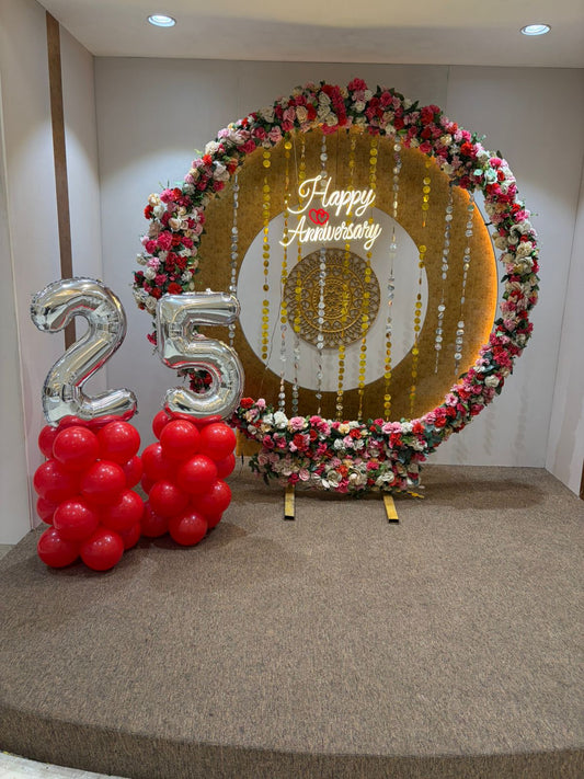 Circular floral ring 25th anniversary backdrop in Jaipur with a neon sign and red balloon pillars by Shava Moments.